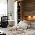 Chambre moderne avec lit escamotable intégré dans un placard en bois, entouré d'étagères avec éclairages encastrés. Un bureau blanc minimaliste est placé à gauche, près d'une grande fenêtre, avec une chaise de bureau noire. Le lit est recouvert d'une couverture en tricot beige et de coussins neutres.