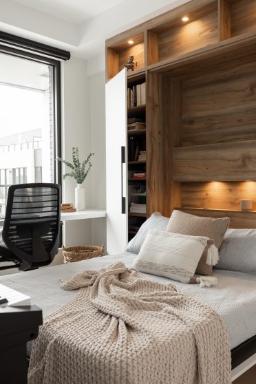 Chambre moderne avec lit escamotable intégré dans un placard en bois, entouré d'étagères avec éclairages encastrés. Un bureau blanc minimaliste est placé à gauche, près d'une grande fenêtre, avec une chaise de bureau noire. Le lit est recouvert d'une couverture en tricot beige et de coussins neutres.