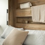 Lit escamotable avec des finitions en bois, intégré à une structure murale avec des étagères. Le lit est recouvert de linge de lit gris clair, accompagné d'un coussin en maille beige. Une couverture assortie est posée sur l'étagère supérieure, ajoutant une touche de confort au design minimaliste et naturel de l'ensemble.