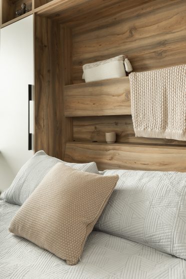 Lit escamotable avec des finitions en bois, intégré à une structure murale avec des étagères. Le lit est recouvert de linge de lit gris clair, accompagné d'un coussin en maille beige. Une couverture assortie est posée sur l'étagère supérieure, ajoutant une touche de confort au design minimaliste et naturel de l'ensemble.