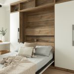 Chambre moderne avec un lit escamotable intégré dans une structure en bois naturel. Le lit, recouvert de linge de lit gris clair et de coussins beige, se replie dans une alcôve entourée d'étagères ouvertes et fermées. Une couverture en maille beige est posée sur le lit, ajoutant une touche de confort. Le sol en bois clair et la décoration minimaliste créent une ambiance épurée et apaisante.