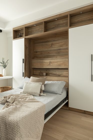 Chambre moderne avec un lit escamotable intégré dans une structure en bois naturel. Le lit, recouvert de linge de lit gris clair et de coussins beige, se replie dans une alcôve entourée d'étagères ouvertes et fermées. Une couverture en maille beige est posée sur le lit, ajoutant une touche de confort. Le sol en bois clair et la décoration minimaliste créent une ambiance épurée et apaisante.