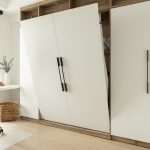 Chambre moderne avec lit escamotable intégré dans un placard en bois, entouré d'étagères avec éclairages encastrés. Un bureau blanc minimaliste est placé à gauche, près d'une grande fenêtre, avec une chaise de bureau noire. Le lit est entrouvert.