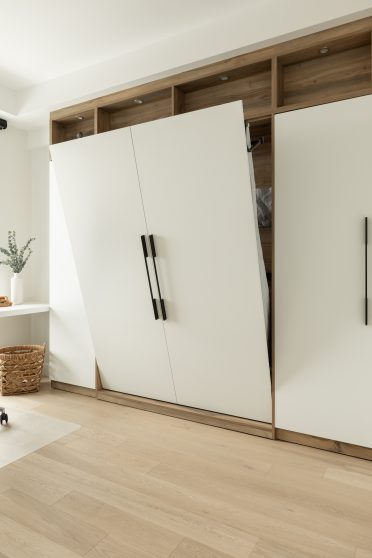 Chambre moderne avec lit escamotable intégré dans un placard en bois, entouré d'étagères avec éclairages encastrés. Un bureau blanc minimaliste est placé à gauche, près d'une grande fenêtre, avec une chaise de bureau noire. Le lit est entrouvert.