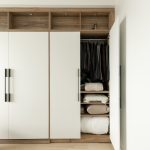 Chambre moderne avec lit escamotable intégré dans un placard en bois, entouré d'étagères avec éclairages encastrés. La porte à droite du lit est entrouverte, montrant une couverture en tricot beige et des coussins neutres.