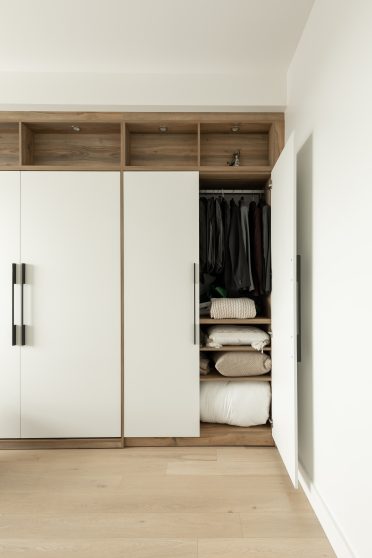 Chambre moderne avec lit escamotable intégré dans un placard en bois, entouré d'étagères avec éclairages encastrés. La porte à droite du lit est entrouverte, montrant une couverture en tricot beige et des coussins neutres.