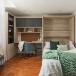 Chambre moderne avec lit escamotable intégré dans un placard en bois, entouré d'étagères et d'un bureau avec éclairages encastrés. Une couverture turquoise est déposée sur le lit donnant une ambiance chaleureuse à l'espace.
