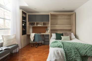 Chambre moderne avec lit escamotable intégré dans un placard en bois, entouré d'étagères et d'un bureau avec éclairages encastrés. Une couverture turquoise est déposée sur le lit donnant une ambiance chaleureuse à l'espace.