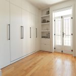 Une chambre minimaliste et lumineuse avec des murs blancs, un lit escamotable rangé. Il y a des étagères intégrées qui contiennent quelques objets décoratifs tels que des livres, des bougies et des plantes. Le sol est en bois clair, et la pièce est baignée de lumière naturelle.