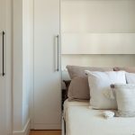 Une chambre minimaliste et lumineuse avec des murs blancs, un lit escamotable avec des draps blancs et une couverture texturée beige. De chaque côté de la porte-fenêtre en verre avec des encadrements noirs, il y a des étagères intégrées qui contiennent quelques objets décoratifs tels que des livres, des bougies et des plantes. Le sol est en bois clair, et la pièce est baignée de lumière naturelle.