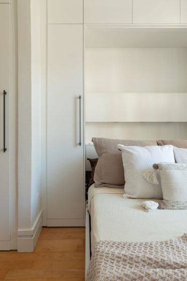 Une chambre minimaliste et lumineuse avec des murs blancs, un lit escamotable avec des draps blancs et une couverture texturée beige. De chaque côté de la porte-fenêtre en verre avec des encadrements noirs, il y a des étagères intégrées qui contiennent quelques objets décoratifs tels que des livres, des bougies et des plantes. Le sol est en bois clair, et la pièce est baignée de lumière naturelle.