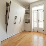 Une chambre minimaliste et lumineuse avec des murs blancs, un lit escamotable rangé. Il y a des étagères intégrées qui contiennent quelques objets décoratifs tels que des livres, des bougies et des plantes. Le sol est en bois clair, et la pièce est baignée de lumière naturelle.