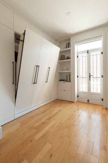 Une chambre minimaliste et lumineuse avec des murs blancs, un lit escamotable rangé. Il y a des étagères intégrées qui contiennent quelques objets décoratifs tels que des livres, des bougies et des plantes. Le sol est en bois clair, et la pièce est baignée de lumière naturelle.