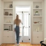 Une chambre minimaliste et lumineuse avec des murs blancs. De chaque côté de la porte-fenêtre en verre avec des encadrements noirs, il y a des étagères intégrées qui contiennent quelques objets décoratifs tels que des livres, des bougies et des plantes. Le sol est en bois clair, et la pièce est baignée de lumière naturelle.