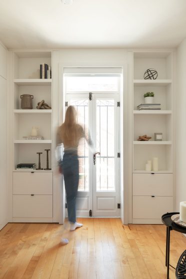 Une chambre minimaliste et lumineuse avec des murs blancs. De chaque côté de la porte-fenêtre en verre avec des encadrements noirs, il y a des étagères intégrées qui contiennent quelques objets décoratifs tels que des livres, des bougies et des plantes. Le sol est en bois clair, et la pièce est baignée de lumière naturelle.