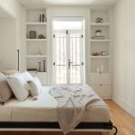 Une chambre minimaliste et lumineuse avec des murs blancs, un lit escamotable avec des draps blancs et une couverture texturée beige. De chaque côté de la porte-fenêtre en verre avec des encadrements noirs, il y a des étagères intégrées qui contiennent quelques objets décoratifs tels que des livres, des bougies et des plantes. Le sol est en bois clair, et la pièce est baignée de lumière naturelle.