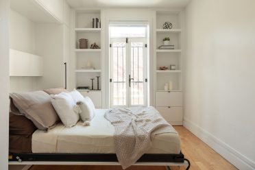 Une chambre minimaliste et lumineuse avec des murs blancs, un lit escamotable avec des draps blancs et une couverture texturée beige. De chaque côté de la porte-fenêtre en verre avec des encadrements noirs, il y a des étagères intégrées qui contiennent quelques objets décoratifs tels que des livres, des bougies et des plantes. Le sol est en bois clair, et la pièce est baignée de lumière naturelle.