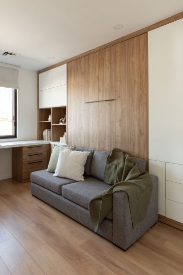 Une chambre moderne et lumineuse avec un canapé-lit escamotable, décoré de coussins beiges et d’une couverture verte au pied. Un petit chien blanc est couché sur le lit. Le mur derrière le lit est revêtu de bois, ajoutant une touche chaleureuse. À gauche, un bureau blanc avec une chaise noire se trouve près d’une grande fenêtre qui laisse entrer la lumière naturelle. Un ensemble d’étagères en bois intégré se situe à l’arrière du bureau, décoré de livres et d’objets. Le sol est en bois clair, complétant le style épuré de la pièce.