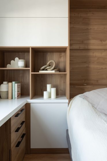 Une chambre moderne et lumineuse avec un lit simple, et blanc. Le mur derrière le lit est revêtu de bois, ajoutant une touche chaleureuse. À gauche, un bureau blanc face à une grande fenêtre qui laisse entrer la lumière naturelle. Un ensemble d’étagères en bois intégré se situe à l’arrière du bureau, décoré de livres et d’objets. 