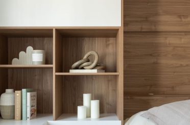 Une chambre moderne et lumineuse avec un lit simple, et blanc. Le mur derrière le lit est revêtu de bois, ajoutant une touche chaleureuse. À gauche, un bureau blanc face à une grande fenêtre qui laisse entrer la lumière naturelle. Un ensemble d’étagères en bois intégré se situe à l’arrière du bureau, décoré de livres et d’objets.