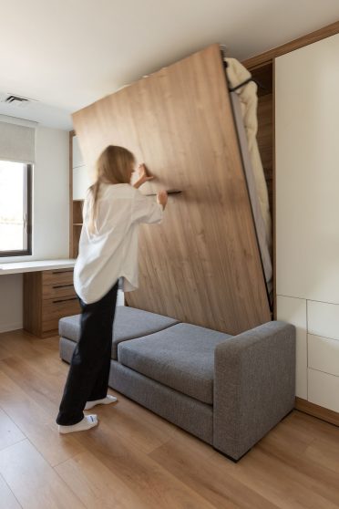 Une chambre moderne et lumineuse avec un canapé-lit escamotable. Une femme aux cheveux blonds, portant un chemisier blanc, range le lit escamotable dans le mur. Le mur derrière le lit est revêtu de bois, ajoutant une touche chaleureuse. À gauche, un bureau blanc avec une chaise noire se trouve près d’une grande fenêtre qui laisse entrer la lumière naturelle. Le sol est en bois clair, complétant le style épuré de la pièce.