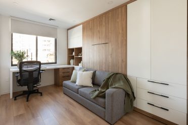 Une chambre moderne et lumineuse avec un canapé-lit escamotable, décoré de coussins beiges et d’une couverture verte au pied. Un petit chien blanc est couché sur le lit. Le mur derrière le lit est revêtu de bois, ajoutant une touche chaleureuse. À gauche, un bureau blanc avec une chaise noire se trouve près d’une grande fenêtre qui laisse entrer la lumière naturelle. Un ensemble d’étagères en bois intégré se situe à l’arrière du bureau, décoré de livres et d’objets. Le sol est en bois clair, complétant le style épuré de la pièce.