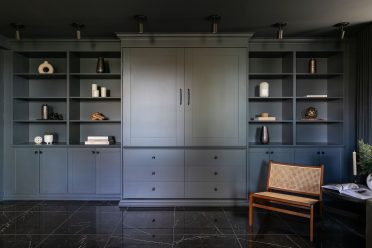 Chambre moderne avec lit escamotable intégré dans un placard en bois bleu, entouré d'étagères. La porte à droite du lit est entrouverte, montrant une couverture en tricot beige et des coussins neutres.