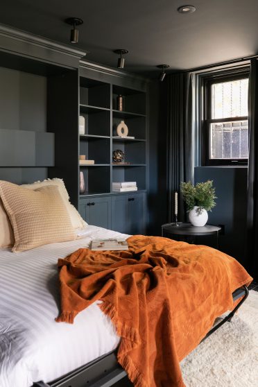 Chambre moderne avec lit escamotable intégré dans un placard en bois bleu, entouré d'étagères. La porte à droite du lit est entrouverte, montrant une couverture en tricot beige et des coussins neutres.