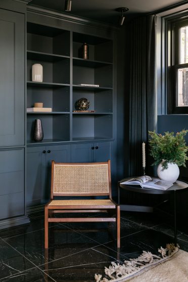 Détail d’un coin salon raffiné avec chaise en rotin moderne, bibliothèque sur mesure en bleu marine profond et table ronde décorée d’un bouquet de feuillage et d’un magazine ouvert.