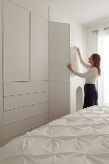 Femme rangeant des vêtements dans un rangement mural sur mesure Limuro, doté de façades blanches minimalistes et d’une niche arrondie intégrée. Un design sobre et pratique qui optimise l’espace de la chambre.