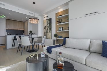 Salon moderne à aire ouverte avec canapé gris, tables basses noires et touches décoratives. À l’arrière, une salle à manger avec table ronde et chaises contemporaines mène à une cuisine minimaliste aux tons blancs et bois, agrémentée d’un luminaire suspendu et d’une œuvre murale.