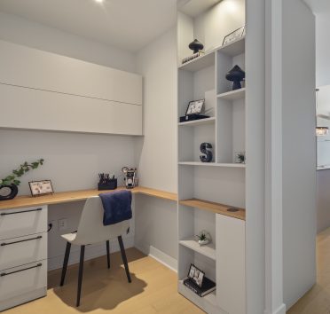 Coin bureau intégré avec bureau en bois et chaise blanche, entouré de rangements ouverts décoratifs. À droite, un couloir mène vers un salon lumineux avec grande fenêtre et vue extérieure.