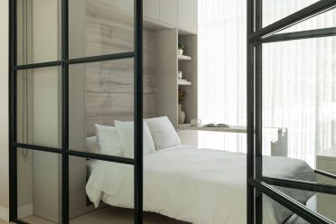 Chambre lumineuse avec lit aux draps blancs, espace bureau intégré et cloison vitrée noire au design contemporain.