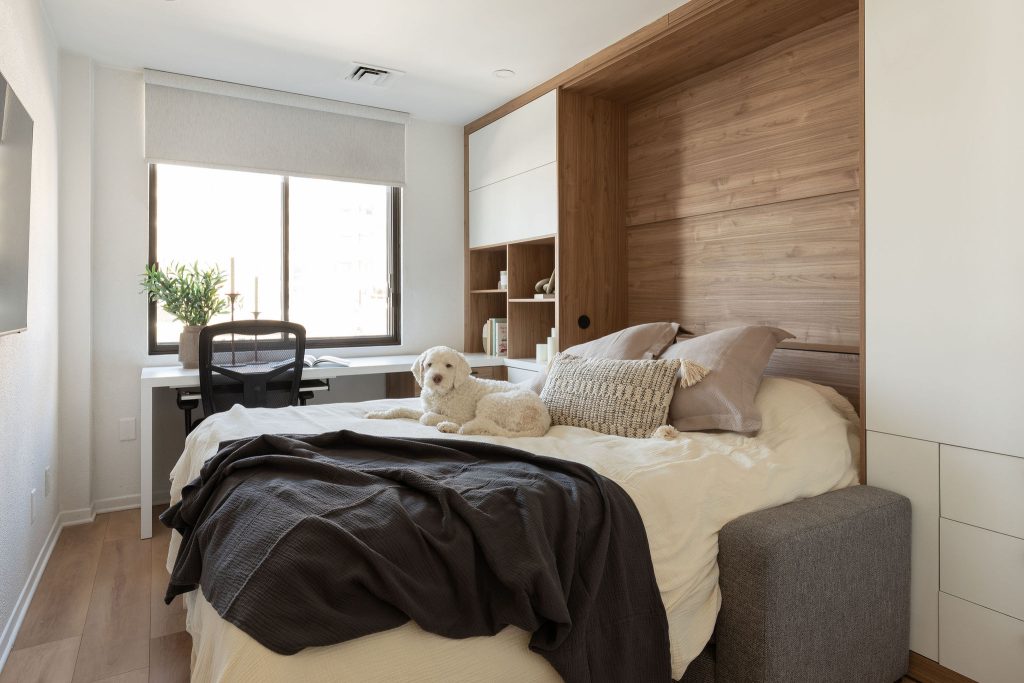Une chambre moderne et lumineuse avec un lit simple, décoré de coussins beiges et d’une couverture noire au pied. Un petit chien blanc est couché sur le lit. Le mur derrière le lit est revêtu de bois, ajoutant une touche chaleureuse. À gauche, un bureau blanc avec une chaise noire se trouve près d’une grande fenêtre qui laisse entrer la lumière naturelle. Un ensemble d’étagères en bois intégré se situe à l’arrière du bureau, décoré de livres et d’objets. Le sol est en bois clair, complétant le style épuré de la pièce.