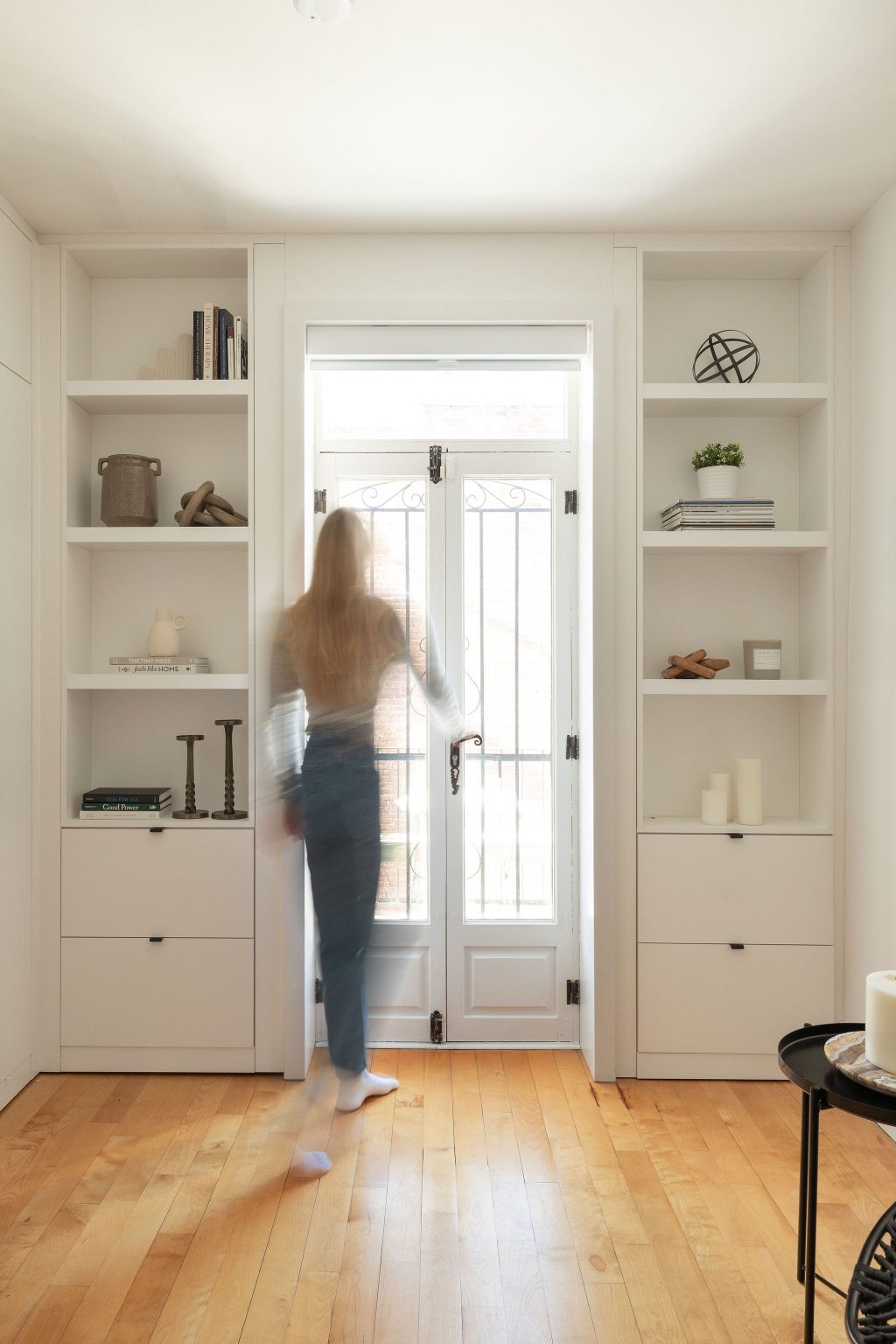 Femme marchant vers une porte-fenêtre dans une pièce épurée aux planchers de bois clair, encadrée de bibliothèques intégrées blanches Limuro.