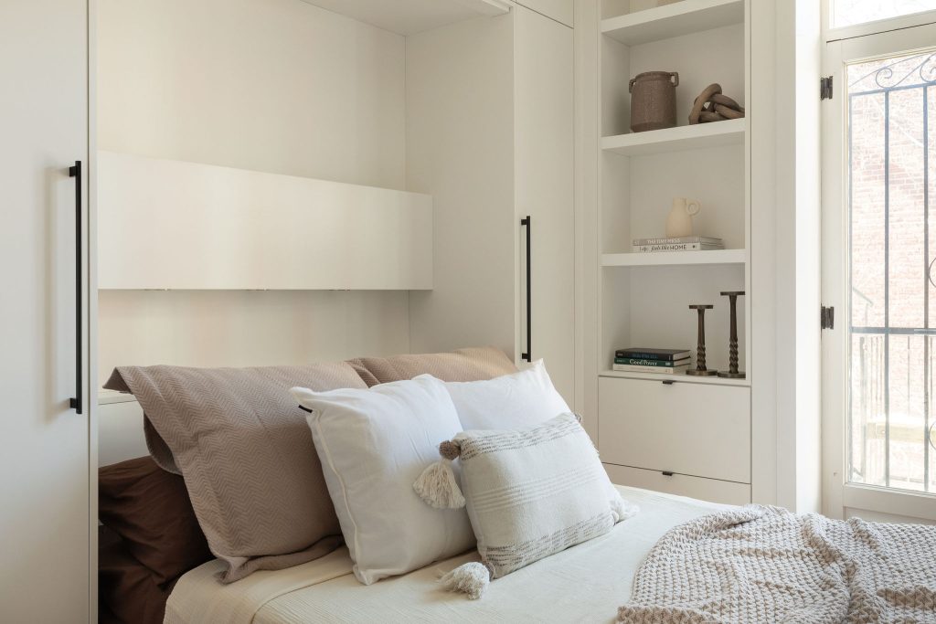 Vue d’ensemble d’une chambre minimaliste avec lit escamotable ouvert, bibliothèques intégrées et portes-fenêtres laissant entrer la lumière naturelle.
