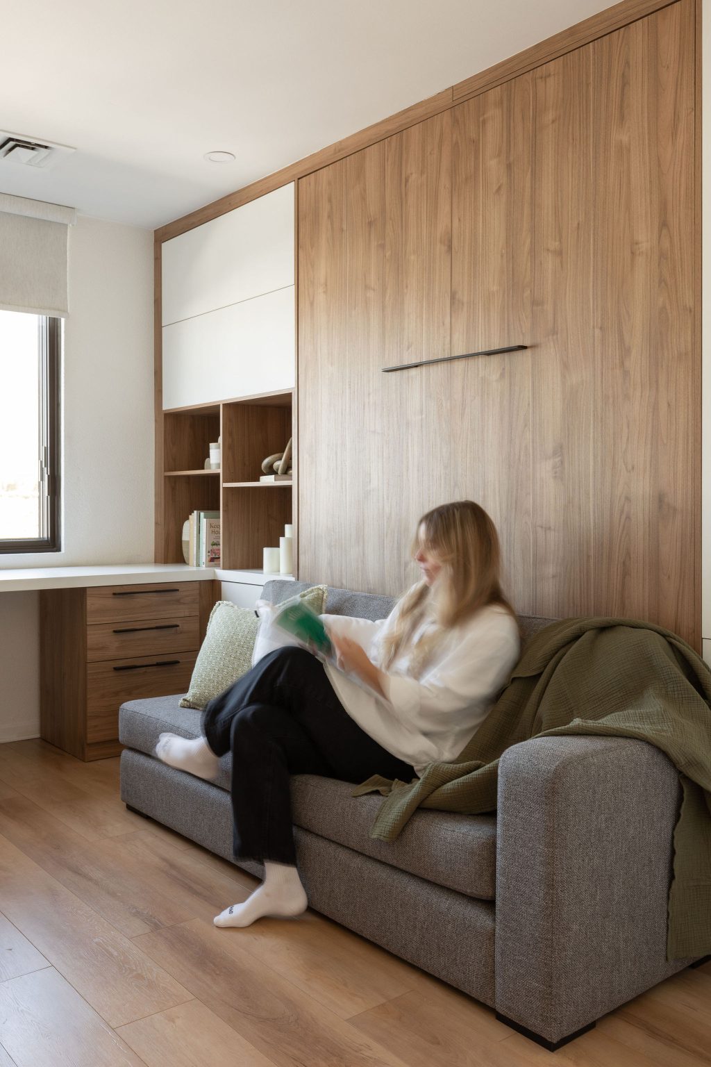 Femme assise sur un sofa gris intégré à un mobilier mural, lisant dans une pièce lumineuse avec rangement en bois clair et bureau adjacent de Limuro