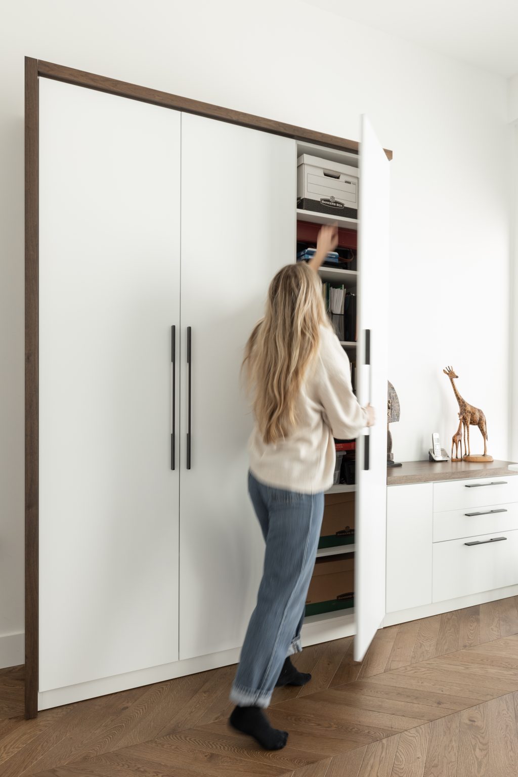 round-image static-image une femme qui ouvre une porte du rangement mural concu sur mesure pour un bureau par Limuro. Sa façade en laque blanche, et son agencement avec le bois le rend intemporel.